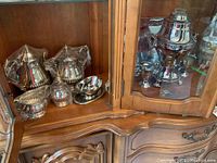 Photo of silver tea and coffee pots, sugar bowl, creamer, and small bowls displayed inside a wooden cabinet.