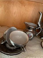 Close-up of demi tasse cups and saucers with silver-tone rims and floral decoration, alongside creamer and sugar bowl.