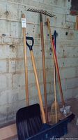 Photo showing shovel, rake, two brooms, and blue pusher leaning against a concrete wall in garage.