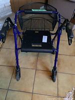 Small blue walker with black seat, mesh storage basket and wheels shown from front on tiled floor