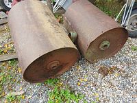 Two heavy metal cylindrical rollers side by side on gravel surface showing light rust and wear
