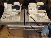 Front view of two beige TEC electronic cash registers side-by-side on a metal storage case showing keypads, receipt printers with paper rolls, and keys on top.