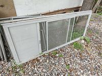 Aluminum screen door with attached sliding glass window panel leaning against a wall outdoors on gravel.