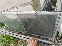 Aluminum framed greenhouse window with two glass panes, held by hand to show size and condition, resting on gravel.