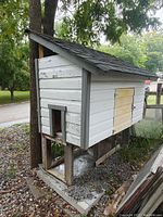 Exterior side view of the white painted wooden chicken coop elevated on wooden legs with small entry door, showing peeling paint and a patched side panel.