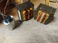 Two rectangular transformers with copper windings and a metal capacitor on wooden surface.