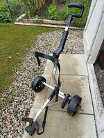 Standing photo of the folding golf bag dolly showing black plastic handle, adjustable straps, and two large wheels on aluminum frame.