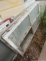 Stack of large wooden windows with chipped white paint and single pane glass divided into four sections, showing weathering and dirt.
