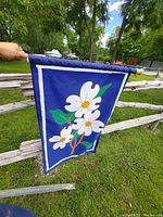 Blue flag with white flowers and green leaves attached to wooden pole hung on a rustic wooden fence in outdoor setting with grass and trees visible.