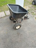 Pull behind spreader on pavement, angled front-left view showing plastic hopper and metal frame with front wheels