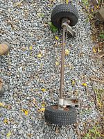 Full view of metal axle with two attached wheels with rubber tires, lying on gravel ground with some grass and leaves.