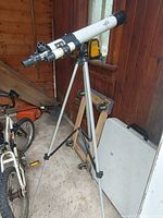 White refractor telescope mounted on adjustable aluminum tripod in wooden room with bicycle and tools in background.