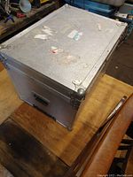 Closed large silver metal travel case placed on a wooden surface showing metal corner protectors and some old label residue on the top and side.
