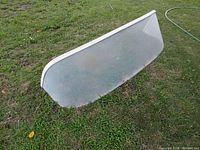 Curved vintage boat window placed on grass, showing front and side profile, illustrating its shape and condition.