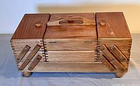 Closed view of wood accordion fold-out sewing box showing natural wood grain finish and two knobs on the sides with one missing knob on top center section.
