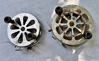Top view of two antique metal fishing reels with dual wooden handles on metal cranks. Shows open circular spokes and patina wear.