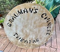 Photo showing front side of large round metal sign with text 'GRAUMAN'S CHINESE THEATRE' painted in black lettering around edge