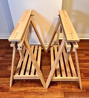 Pair of wooden sawhorses shown from an angle, open and standing, displaying structure, top beams, and lower slatted shelves.