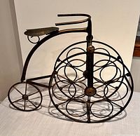 Side view of metal bicycle wine bottle holder showing large front wheel with circular loops to hold wine bottles and smaller rear wheel.