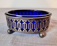 Side view showing the sterling silver holder with intricate cutout pattern and cobalt blue glass insert inside, resting on three ball feet.