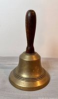Full front view of vintage brass school bell with wood handle on flat surface.