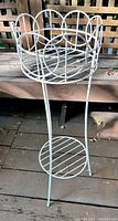 Full-view photo of the white painted vintage cast iron metal plant stand with scalloped circular top and lower shelf set on wooden deck.
