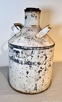 Full view of the vintage metal jug showing spouts, handle, and weathered white paint with black lettering.