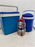 Two blue beverage coolers and one clear plastic sun tea maker with a cow design and spigot. The coolers have white handles and lids, the sun tea maker has a red lid and handle.