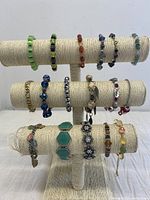 Front view of assortment of 17 colorful costume summer bracelets displayed on a beige bracelet stand. Bracelets are made of beads, crystals, hematite, and charms in various colors and styles.