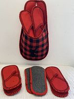 Hanging red and black plaid fabric bag with black deer silhouette containing two pairs of slippers, and three slippers laid below front facing.