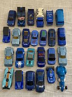 Top down photo showing 25 blue metal toy cars arranged in 5 rows, including race cars, trucks, and sedans with various decals and paint.
