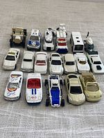 Top-down view of 19 white and beige diecast toy vehicles, arranged in 3 rows showing various types from cars to helicopter with some vehicles having doors open