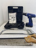 Power Smith 18V cordless drill, black plastic carrying case, instruction manual, back saw with wooden handle, and hack saw arranged on table.