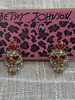Pair of Betsey Johnson skull-shaped gold tone pierced earrings with multicoloured faceted rhinestones and pink enamel rose, set against a leopard print Betsey Johnson box.