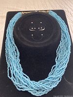 Front view of multi-strand turquoise seed bead necklace displayed on black stand.