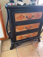 Full front view of the wooden cabinet showing three drawers with diamond-shaped carved patterns and metal handles, against a tiled floor and a wooden-framed mirror beside it.