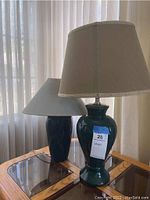 Two ceramic table lamps on a glass and wood table by a curtained window, the taller lamp has a beige shade marked 23 inches tall
