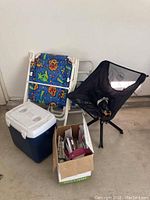 Two CLIQ camping chairs (one visible fully in black mesh), two metal-framed blue tropical patterned fabric beach chairs, blue-and-white Coleman cooler, and a box of plastic plates and disposable cutlery.