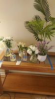 Photo showing six different potted faux flower arrangements on a wooden table with two soap dispensers.
