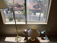 Photo showing the brass standing floor lamp and three table lamps placed on the floor near a window, with a view outside.