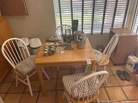 Square wooden kitchen table with four white spindle-back chairs and checkered seat cushions with items on the table not included for sale.
