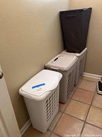 View of three cream plastic laundry baskets with ventilation holes and white lids, lined up against a wall next to a tall dark woven hamper