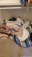 Pile of assorted bed sheets and blankets in beige, white, and striped patterns stacked on the floor of a closet.
