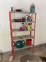 Metal shelving unit with red frame and 5 scratched white shelves, holding automotive and household supplies, gardening watering can, and other small items.