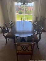 Mid century dining table with six chairs visible around it, showing the oval wooden table top and cushioned chairs in a light fabric, with window and curtain background.
