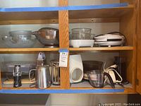 Various clear glass mixing bowls on the top shelf along with metal colander and white waffle maker next to them. Bottom shelf contains electric coffee grinder, metal measuring cups, and other kitchen tools.