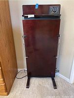 Front view of the tall wooden pants press standing on carpeted floor showing the closed press with metal side handles and legs.