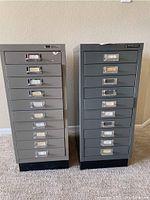 Front view of two small filing cabinets with 10 drawers each, showing metal pull handles with label holders, signs of scratches and wear on cabinet surfaces.