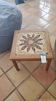 Full view of square wooden side table on tiled floor showing tiled patterned top and wooden legs.