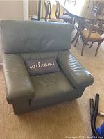 Green upholstered armchair seen from the front, showing padded armrests and backrest, with a "welcome" pillow in the seat.